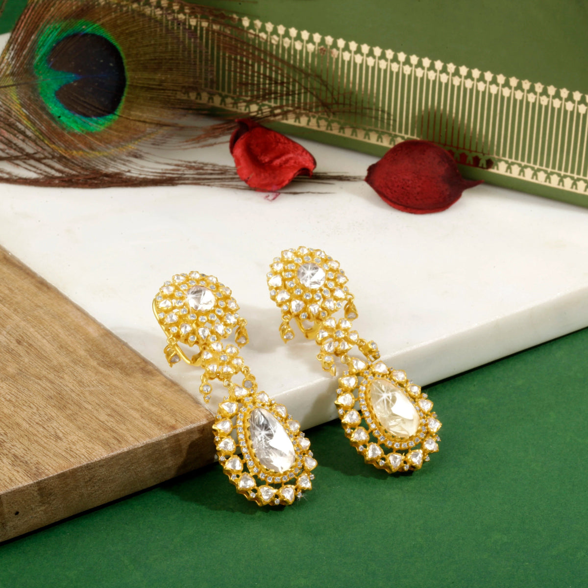 A pair of silver earrings with gold accents and sparkling details, displayed on a white surface with a peacock feather and red petal in the background.