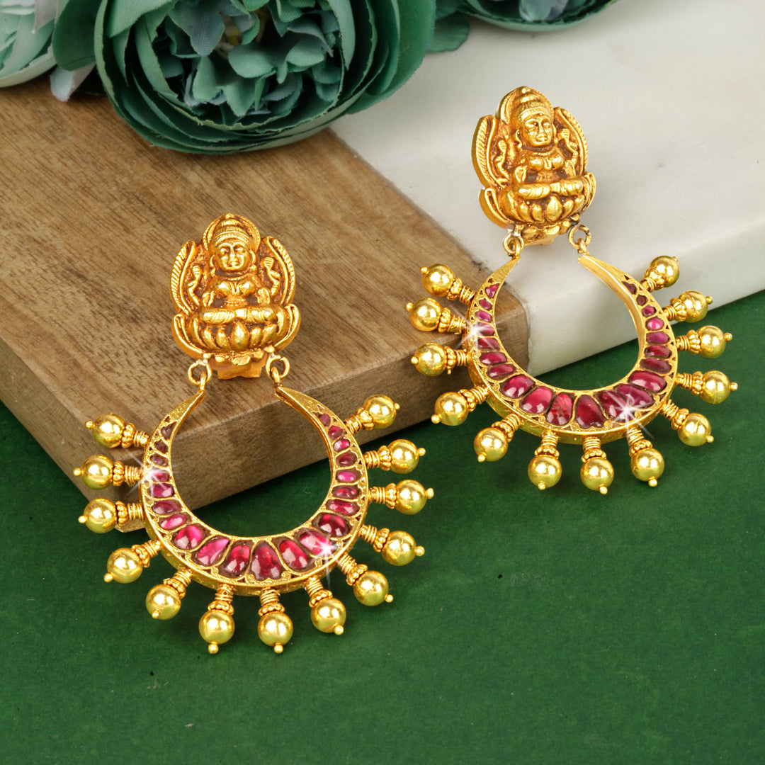 A pair of gold-plated silver earrings with red stone inlays, displayed on a wooden surface with a floral background.