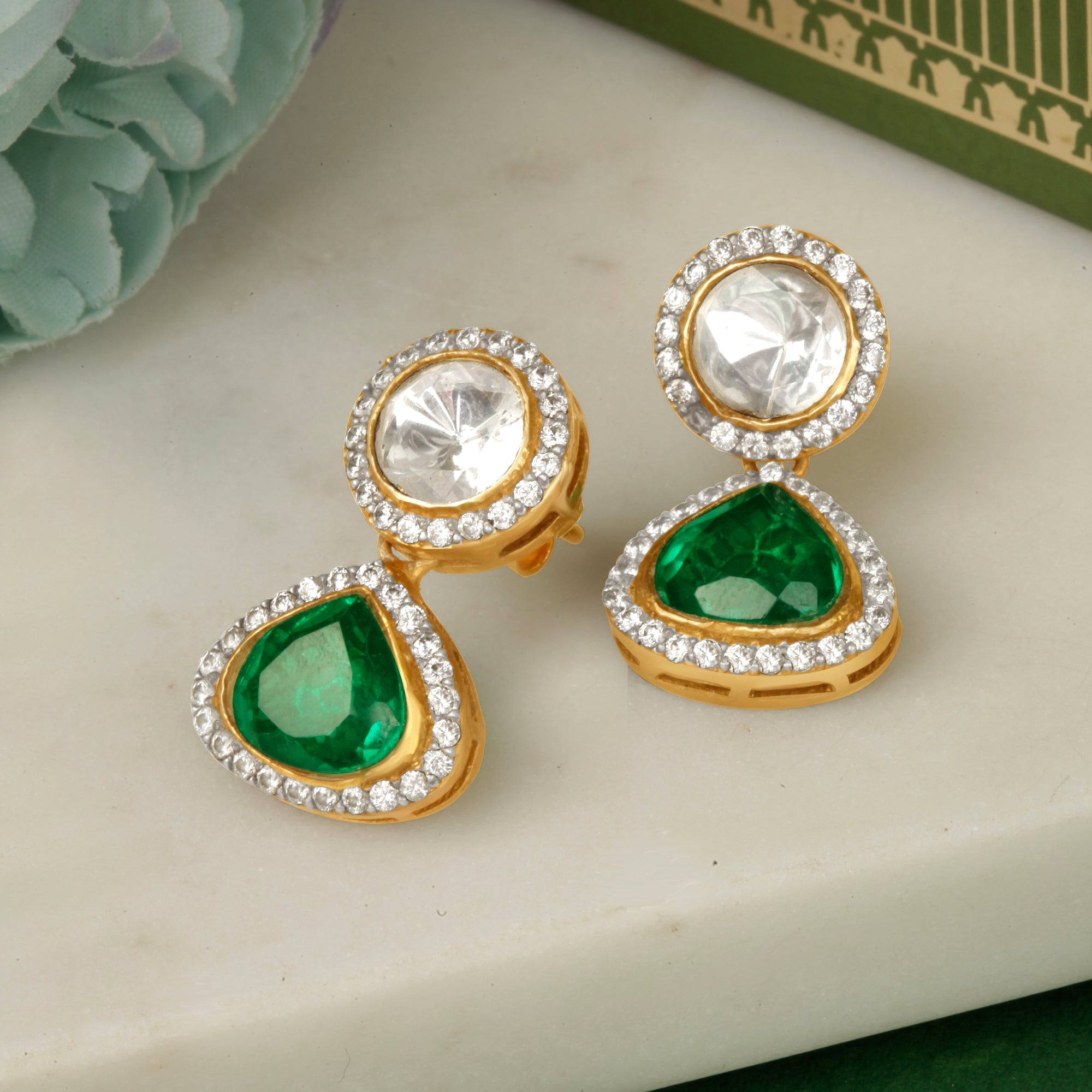A pair of silver earrings with green stones and white accent stones, displayed on a marble surface with a green fabric backdrop.