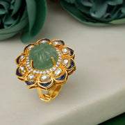 Silver Gold plated ring with green gemstone and blue accents on a white surface with green flowers in the background